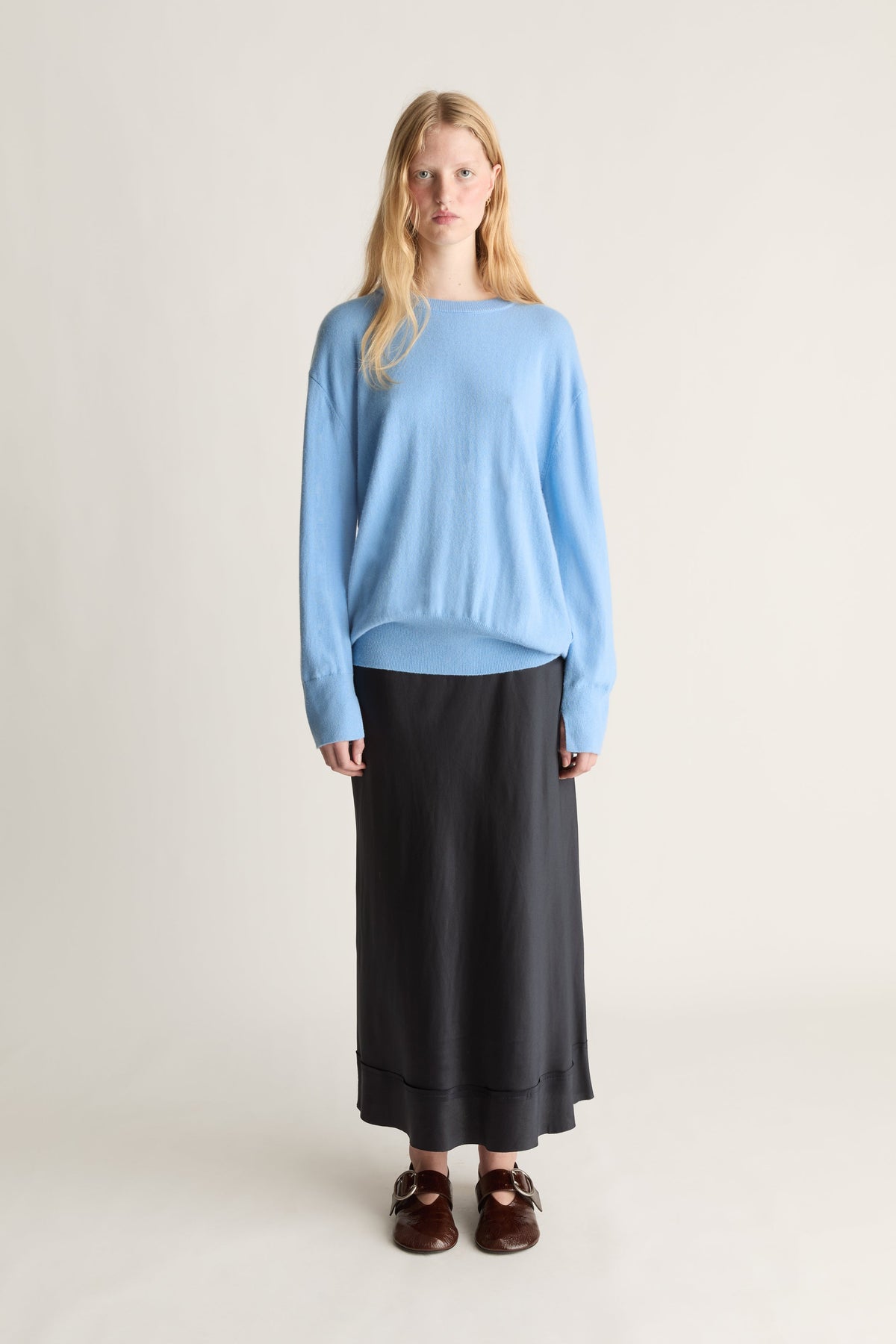 A woman stands against a plain light beige studio background wearing a light blue long-sleeve knit top, a long black skirt, and dark brown loafers.