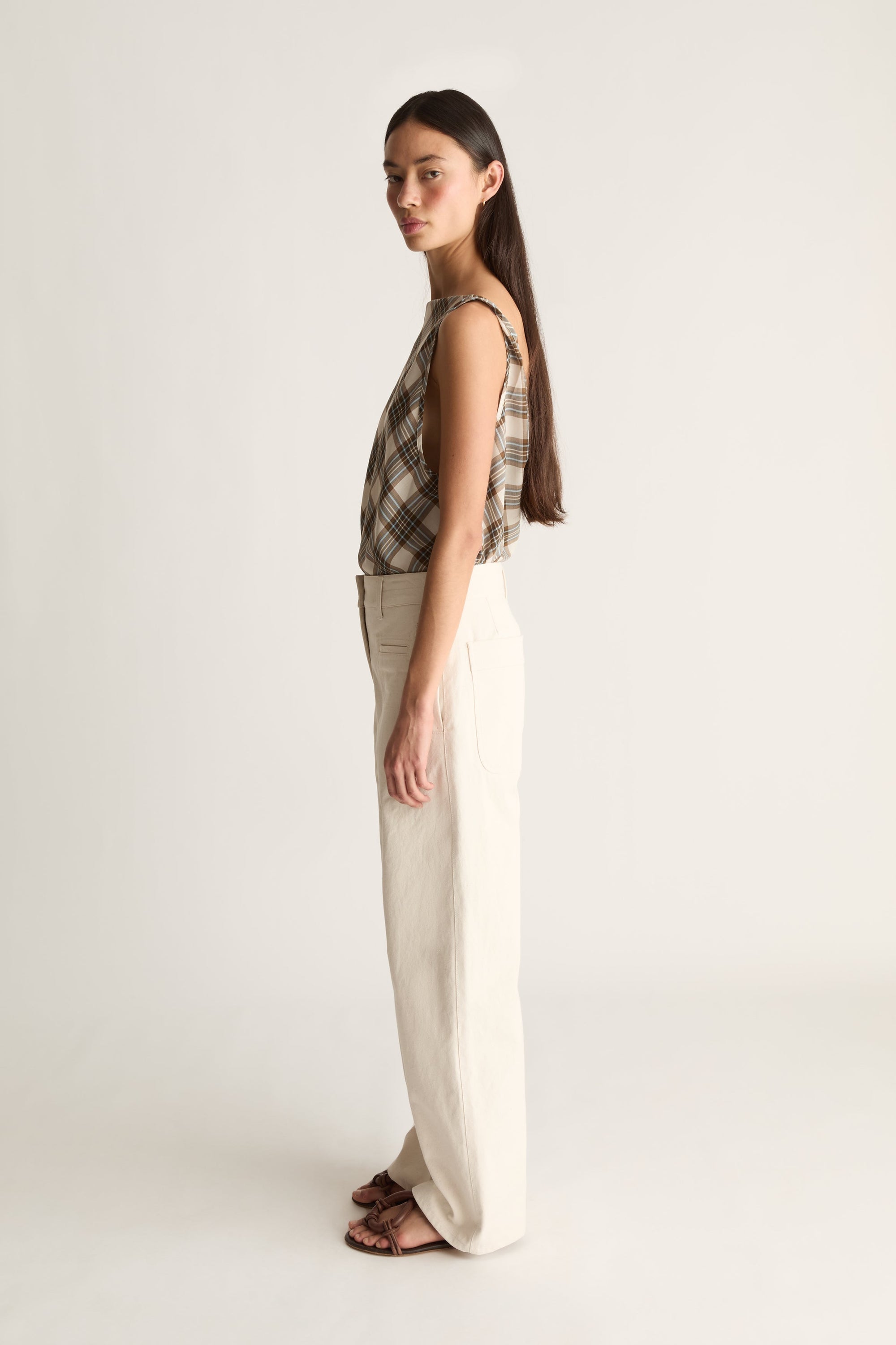 A woman stands in profile wearing a sleeveless plaid top in beige and brown tones with wide-leg cream trousers and brown sandals, photographed against a plain light gray studio background.