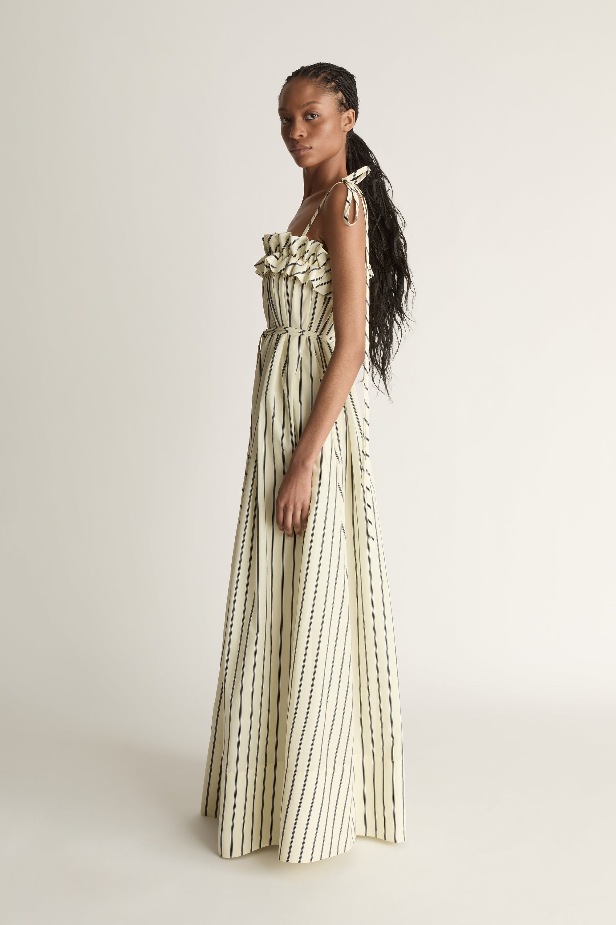 A side view of a woman wearing a lemon yellow and black striped cotton dress with a waist tie on a white background