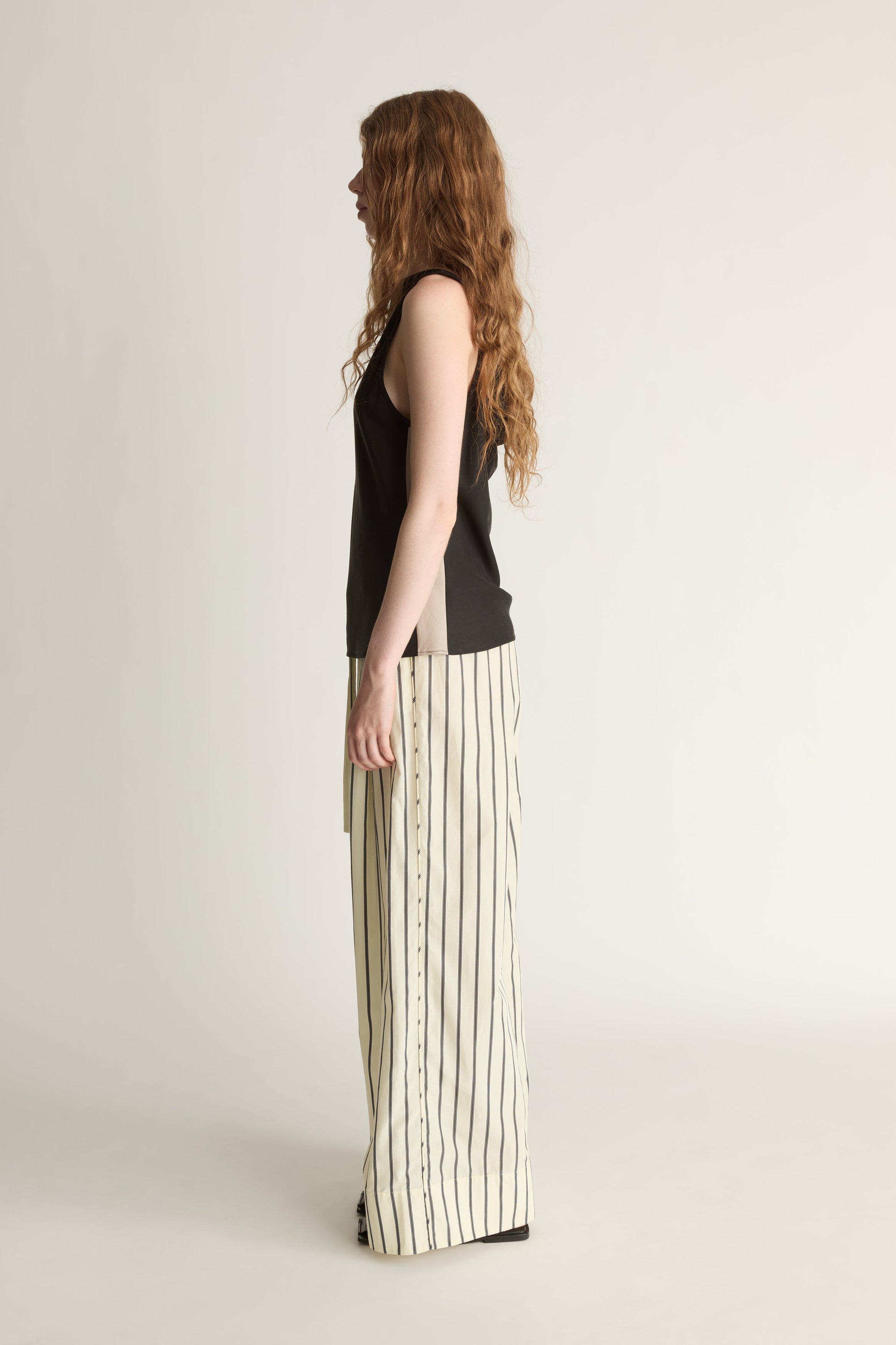 A side view of a woman wearing a pair of Yellow and black striped pants and a black bias cut top on a white background.