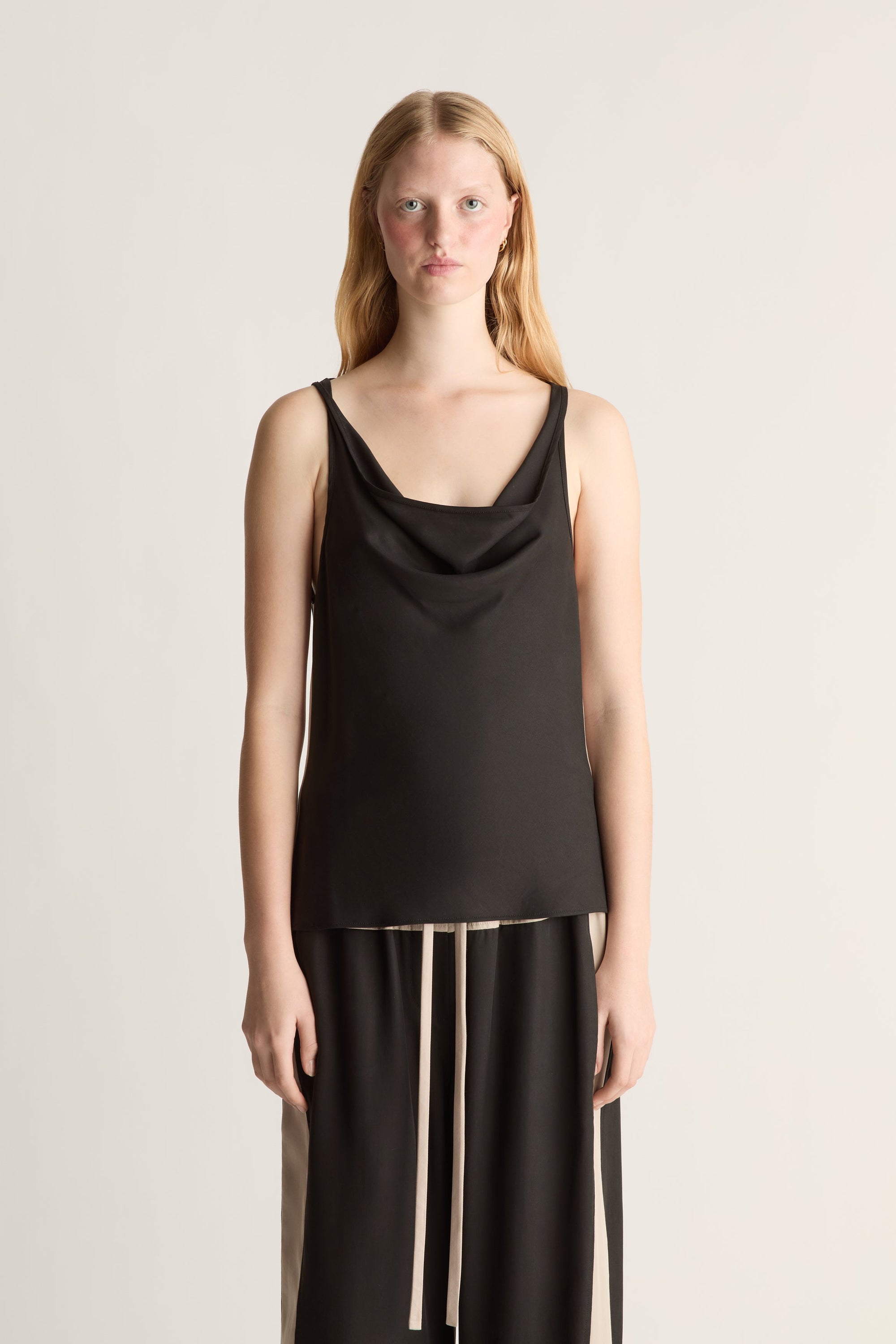 A woman wearing a black cowl neck sleeveless shirt and black trousers on a white background