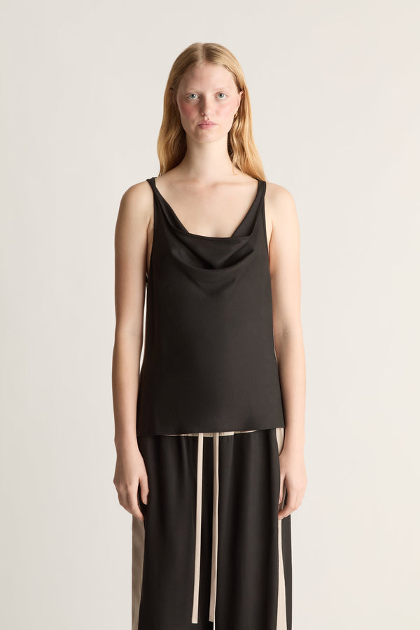 A woman wearing a black cowl neck sleeveless shirt and black trousers on a white background