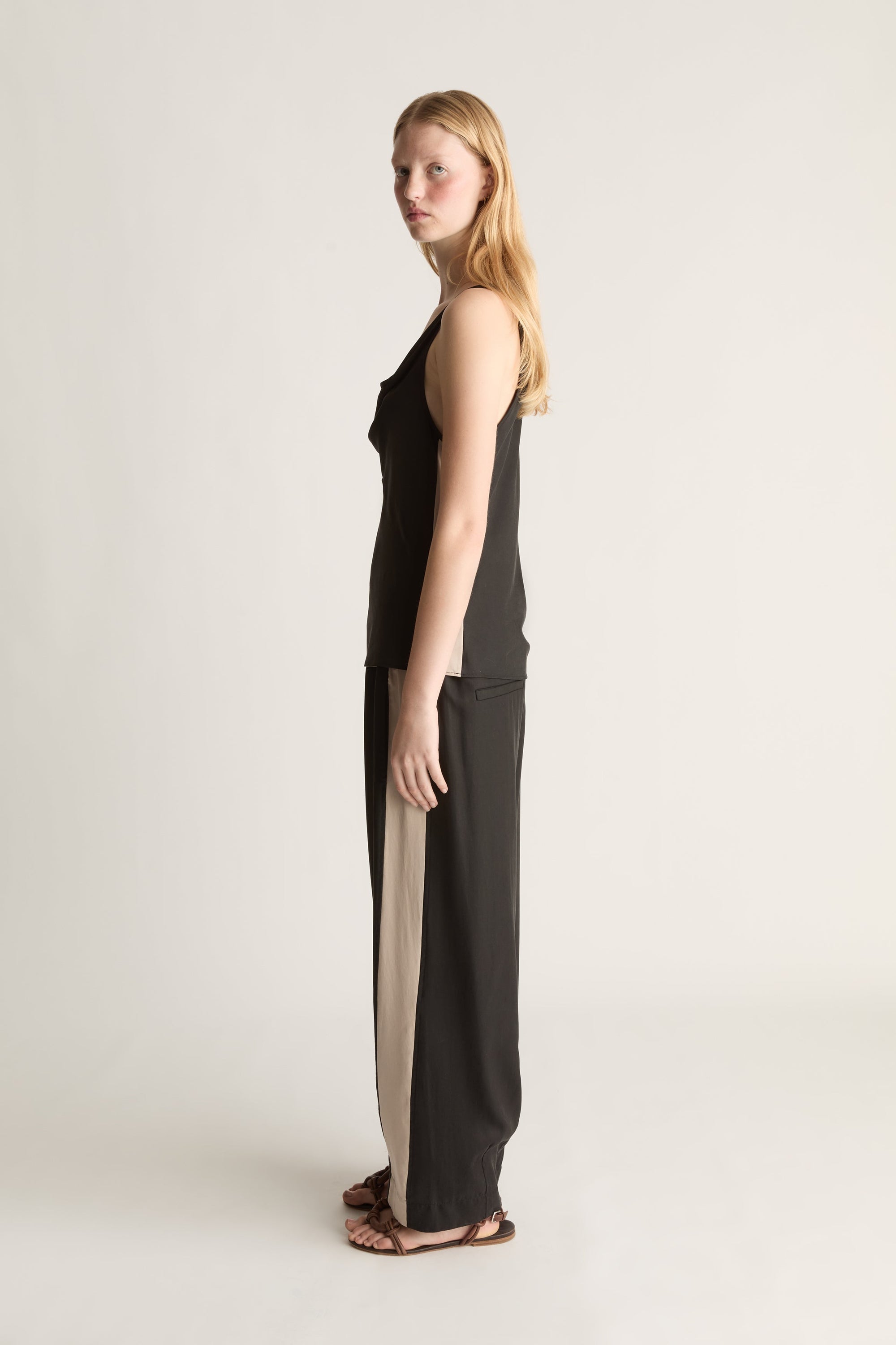 Side view of a woman wearing a sleeveless black blouse and a pair of black trousers with a white stripe down the side on a white background.