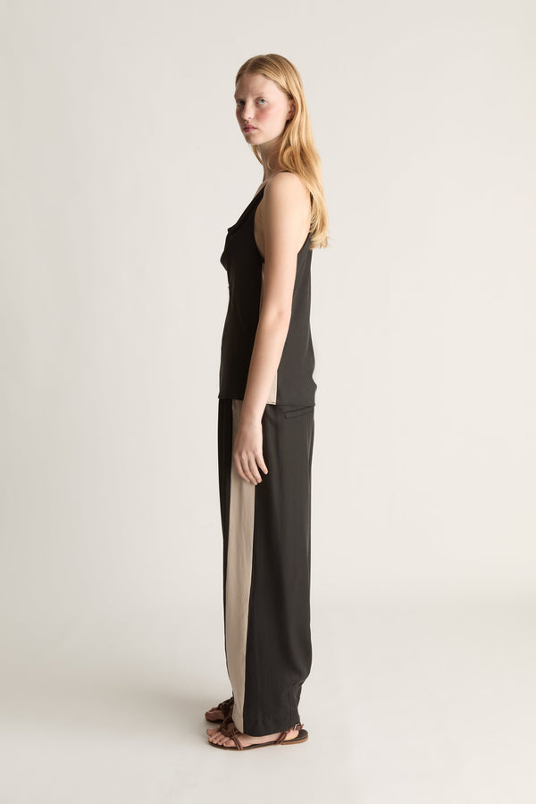 Side view of a woman wearing a sleeveless black blouse and a pair of black trousers with a white stripe down the side on a white background.