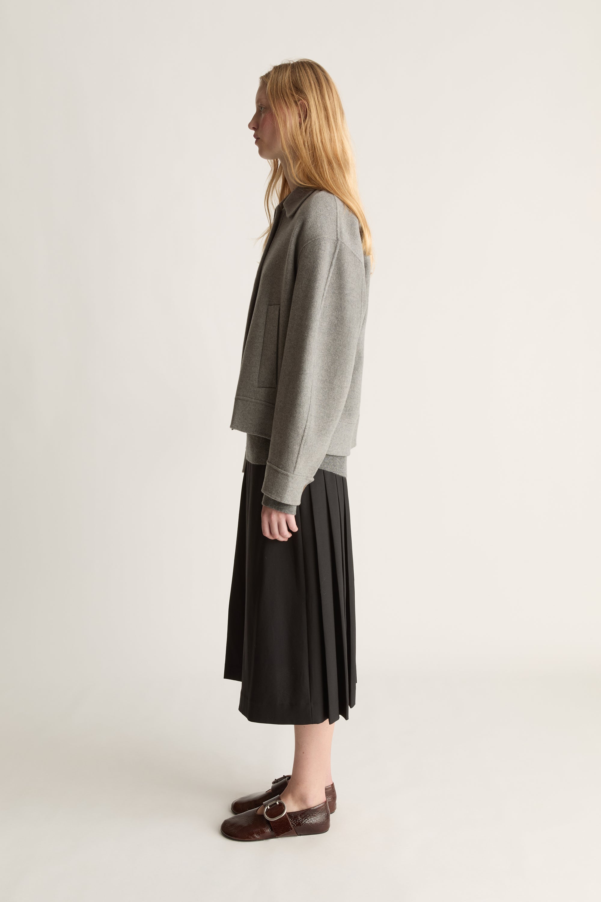 Side view of a woman wearing a grey cashmere jacket with a black kilt skirt and brown shoes against a white studio background. 