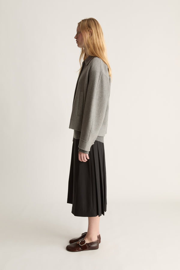 Side view of a woman wearing a grey cashmere jacket with a black kilt skirt and brown shoes against a white studio background. 