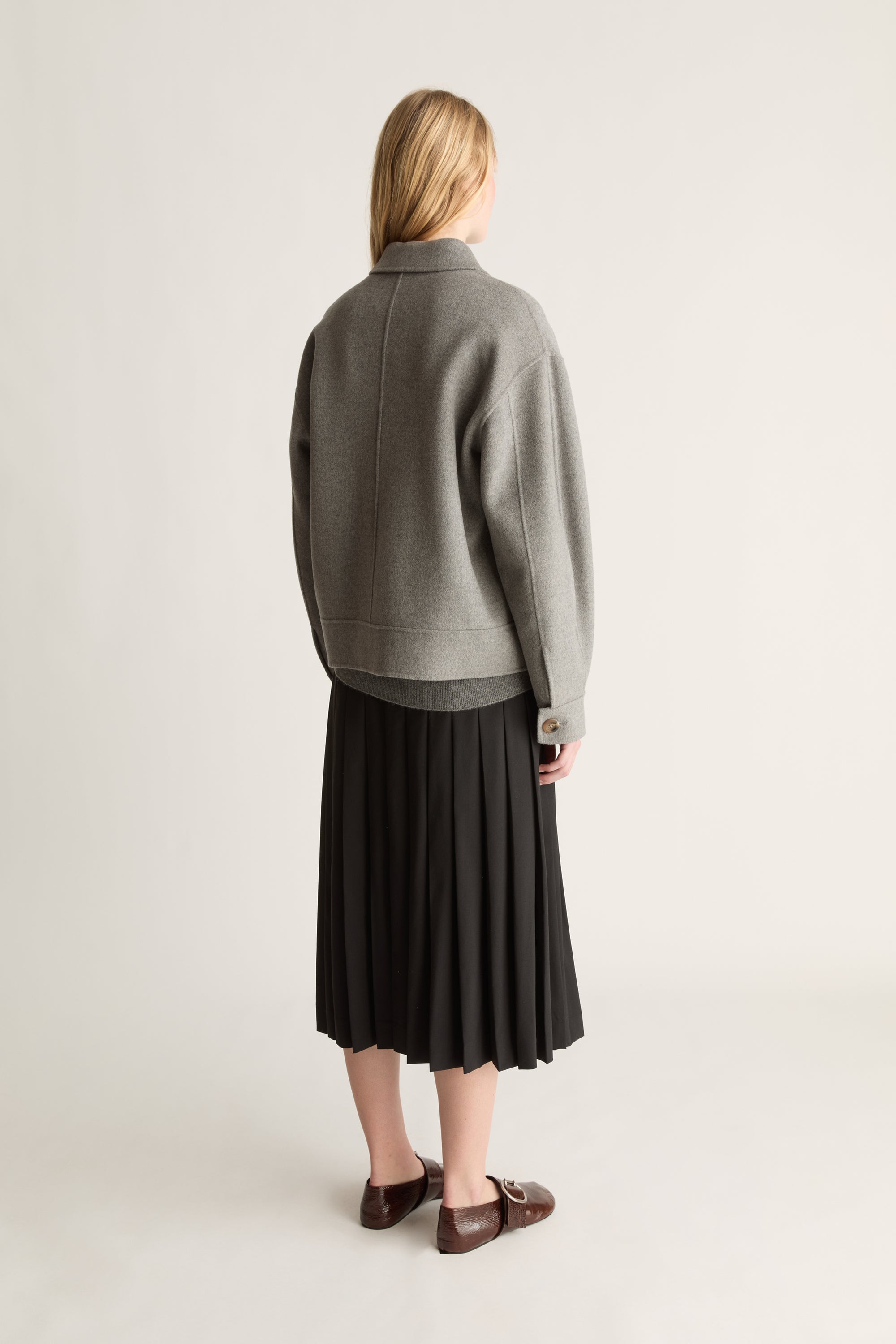 Back view of a woman wearing a grey jacket, a black wool kilt skirt and brown shoes against a white studio background..