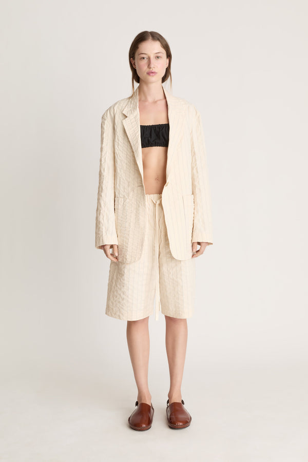 Woman wearing a cream blazer and shorts with a black bralette underneath on a white background.