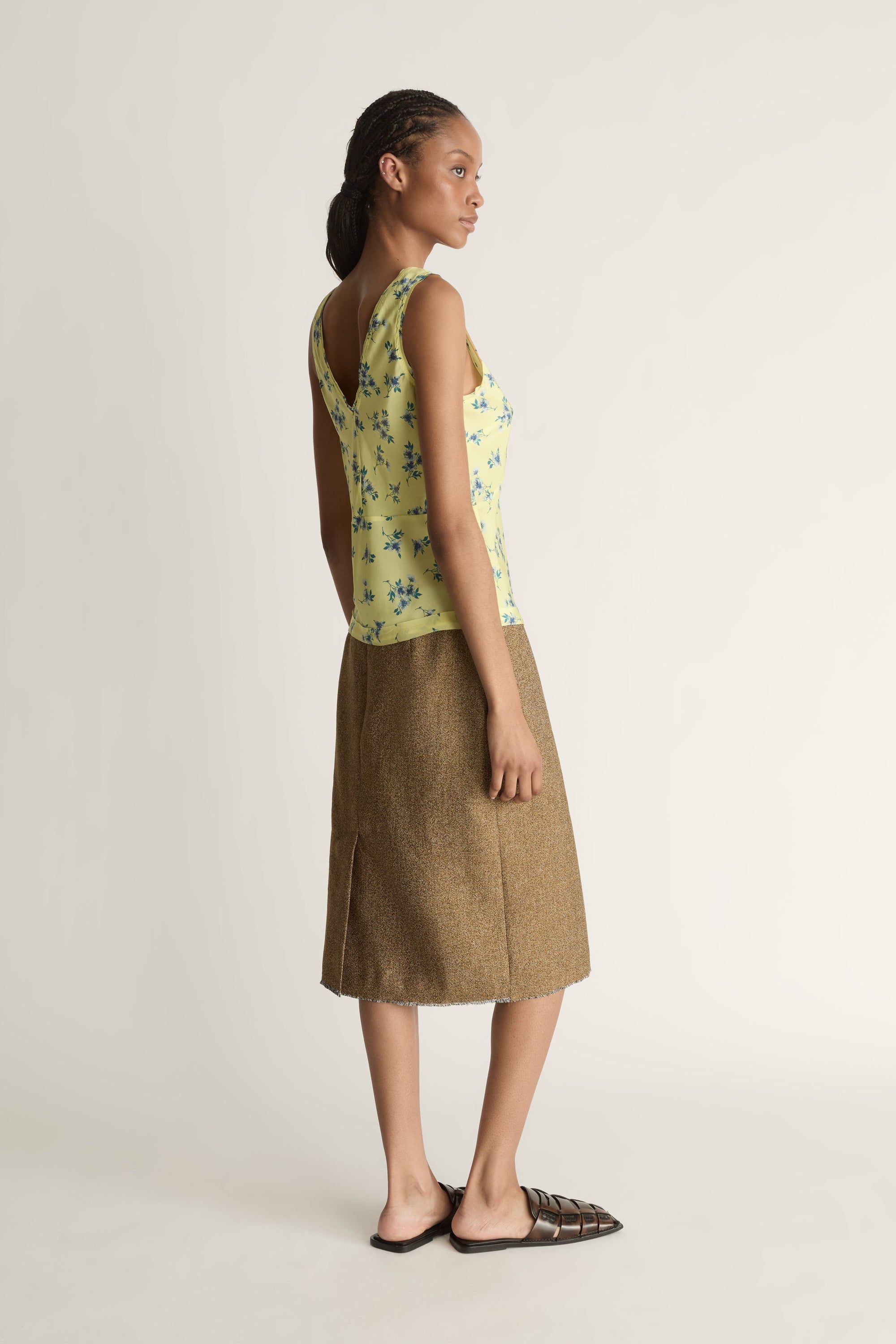 Side view image of a woman wearing a yellow V-neck blouse with blue flowers on it and a brown skirt on a white background.