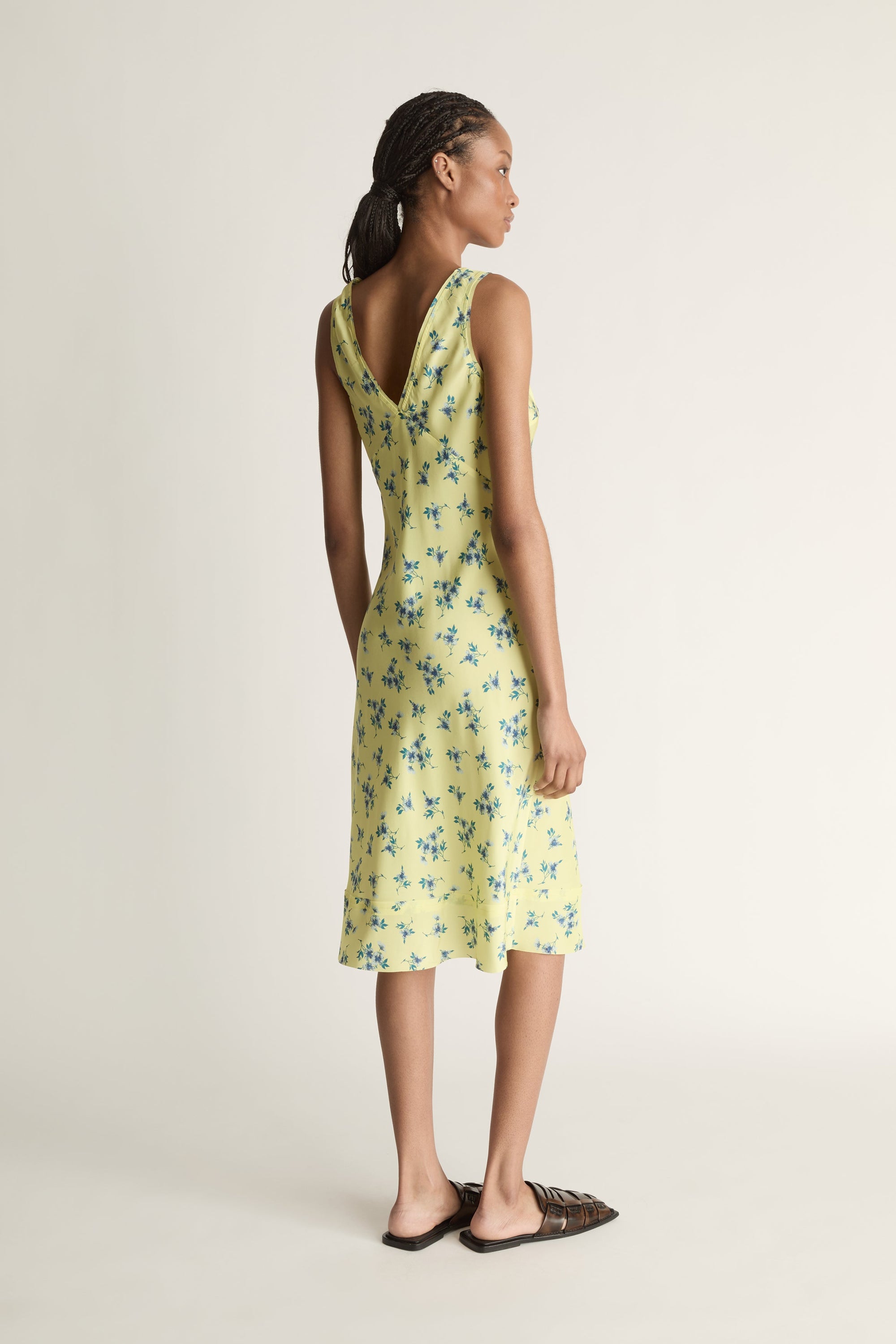 Back view image of a woman wearing  the Maisie Silk slip dress in a lemon yellow fabric that features a blue floral print. 
