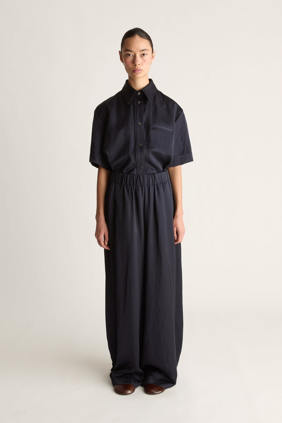 Front view of a woman wearing a navy short-sleeved shirt and elasticated wide-leg trousers, shot against a white studio background.