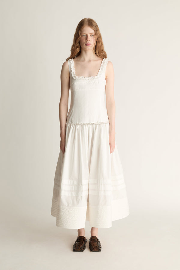 A woman stands against a plain light beige studio background wearing a sleeveless cream dress with a square neckline, delicate lace trim at the shoulders and waist, and a tiered skirt. She is wearing dark brown loafers.