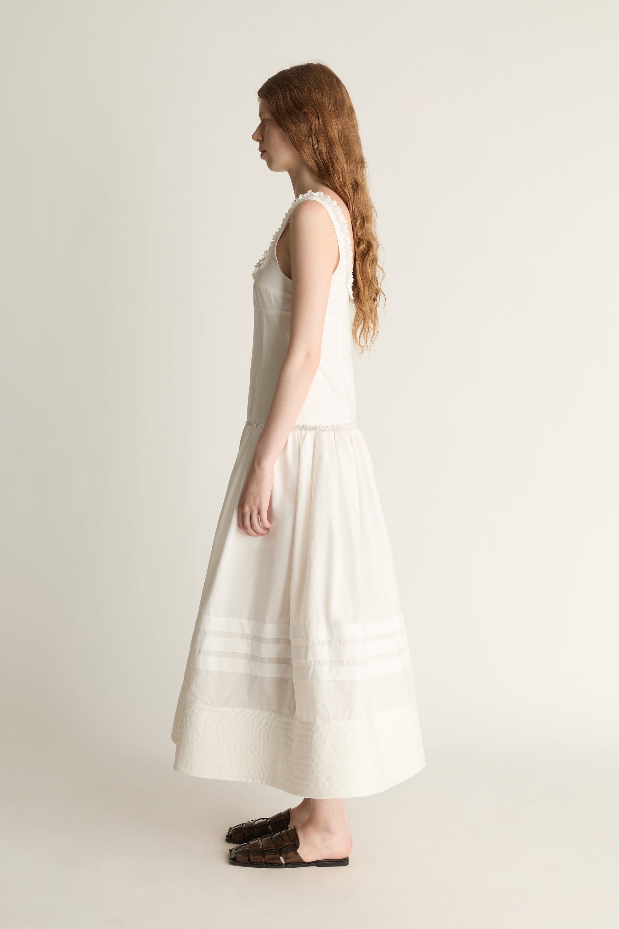 A woman in profile wearing a sleeveless cream-colored midi dress with horizontal embroidered stripes near the hem, paired with dark brown flat shoes, set against a plain light beige studio background.