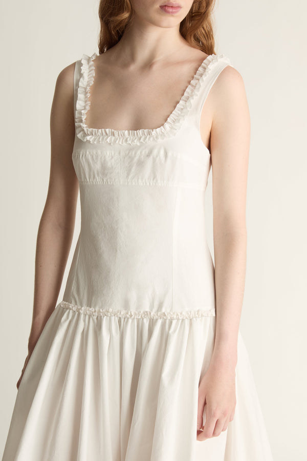 Zoom in on a woman wearing a sleeveless cream-colored dress with ruffled trim along the scoop neckline and waist, shown against a plain light background.