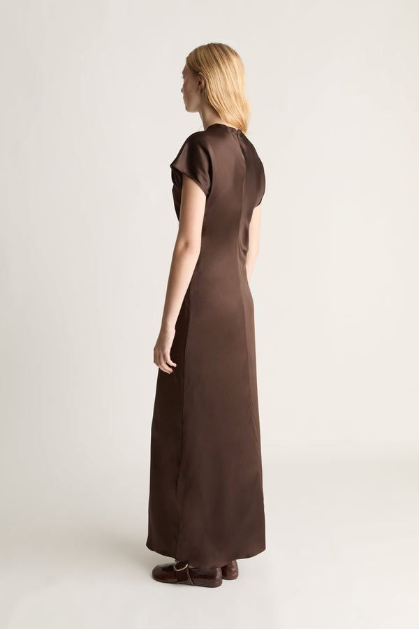 Back view of a woman wearing a brown short sleeve full length dress with brown shoes against a white background.