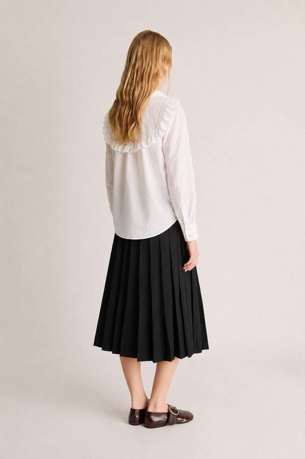 A woman shown from the back wearing a white long-sleeve blouse with a ruffled collar and a black pleated midi skirt, paired with dark brown loafers, against a plain light beige studio background.