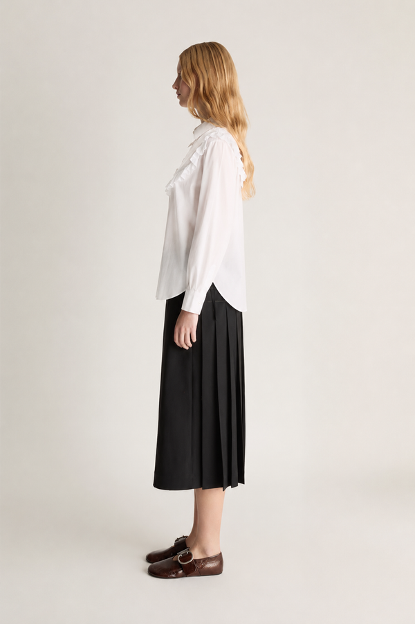 A woman stands in profile wearing a white long-sleeve blouse with ruffled detailing on the shoulders and a black pleated midi skirt, paired with dark brown loafers, against a plain light gray studio background.